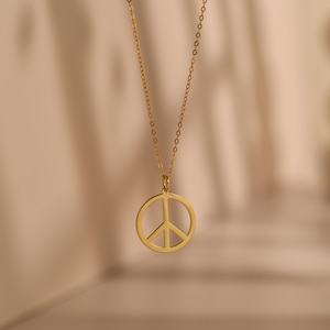 May include: A gold-colored necklace featuring a peace symbol pendant. The pendant is a circular design with a vertical and two diagonal lines, representing peace. The necklace chain is delicate and hangs against a soft, neutral background.