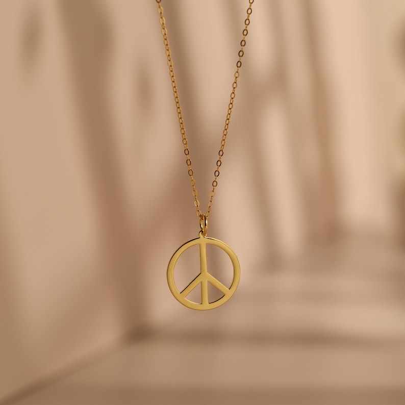 May include: A gold-colored necklace featuring a peace symbol pendant. The pendant is a circular design with a vertical and two diagonal lines, representing peace. The necklace chain is delicate and hangs against a soft, neutral background.