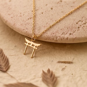 Könnte beinhalten: Eine goldene Halskette mit einem kleinen Torii-Tor-Anhänger. Die Kette liegt auf einer hellbraunen Oberfläche.