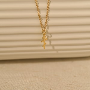 May include: A gold-colored necklace with a pendant in the shape of a cross. The pendant has a unique design with three horizontal bars. The chain is delicate and the necklace is displayed against a neutral background.