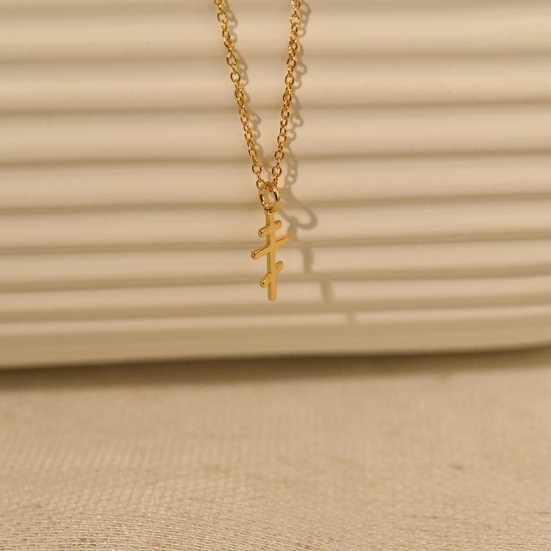 May include: A gold-colored necklace with a pendant in the shape of a cross. The pendant has a unique design with three horizontal bars. The chain is delicate and the necklace is displayed against a neutral background.