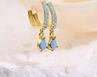 May include: Gold hoop earrings with a light blue teardrop charm dangling from each hoop. The hoops are decorated with a row of light blue stones.