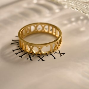 May include: A gold-toned ring with a Roman numeral design. The ring is made of metal and has a simple, elegant style.