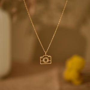 May include: A gold chain necklace with a camera pendant. The camera is a simple outline with a circle for the lens.