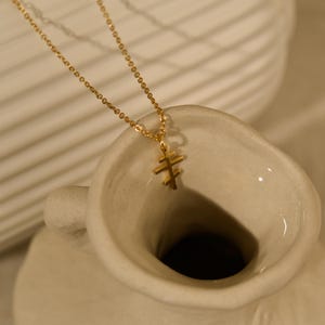 May include: A gold-colored necklace with a double-barred cross pendant. The necklace chain is delicate, and the pendant is a simple, elegant design. The necklace is displayed against a neutral background, highlighting its minimalist aesthetic.