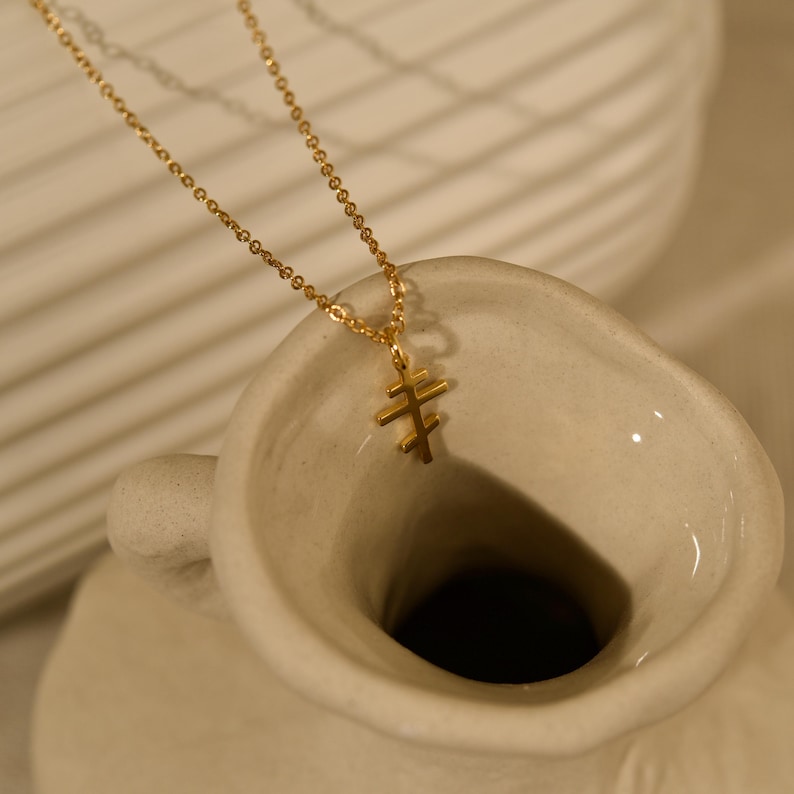 May include: A gold-colored necklace with a double-barred cross pendant. The necklace chain is delicate, and the pendant is a simple, elegant design. The necklace is displayed against a neutral background, highlighting its minimalist aesthetic.