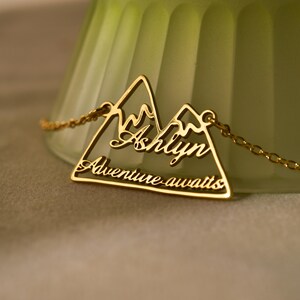 以下が含まれることがあります： 山脈のペンダントが付いたゴールドネックレス。ペンダントには「Ashlyn」という名前と「冒険が待っている」というテキストが書かれています。
