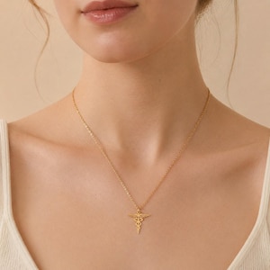 Può includere: Una collana dorata con un ciondolo Caduceo, simbolo della medicina. Il ciondolo è appeso a una catena sottile. La collana è indossata sulla pelle di una persona, con uno sfondo morbido e neutro.
