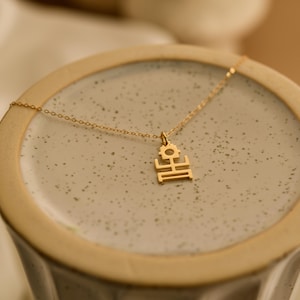 Puede incluir: Un delicado collar dorado con un colgante con un diseño geométrico. El colgante está suspendido de una fina cadena. El collar se exhibe sobre una superficie de cerámica gris claro, creando una estética minimalista.