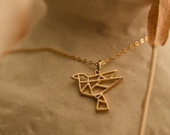 May include: A gold-colored necklace featuring a geometric bird pendant. The bird design is made of interconnected lines, creating a modern, minimalist aesthetic. The necklace is displayed against a neutral background, highlighting the jewelry's design.