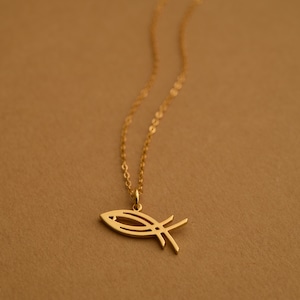 Puede incluir: Collar dorado con un colgante de pez estilizado. El colgante tiene un diseño minimalista con líneas limpias. La cadena es delicada y el fondo es de un marrón cálido.