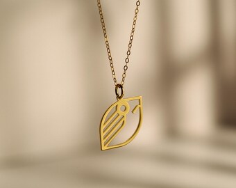 Pode incluir: Um colar com pingente dourado com um design em forma de folha. O pingente tem um design recortado com três linhas paralelas e um detalhe circular. A corrente do colar também é dourada e pende sobre um fundo neutro.