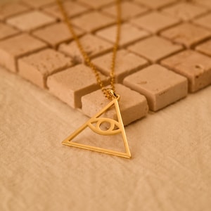 May include: A gold-toned necklace featuring a triangular pendant with an open-work eye design in the centre. The pendant is suspended from a delicate gold-toned chain, showcasing a polished metallic finish and a geometric, symmetrical aesthetic.
