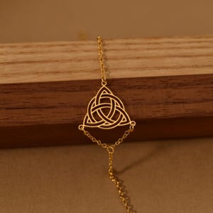 May include: A gold-colored necklace featuring a Celtic knot pendant. The pendant is a triangular shape with an intricate interwoven design. The necklace has a delicate chain and is displayed against a wooden background.