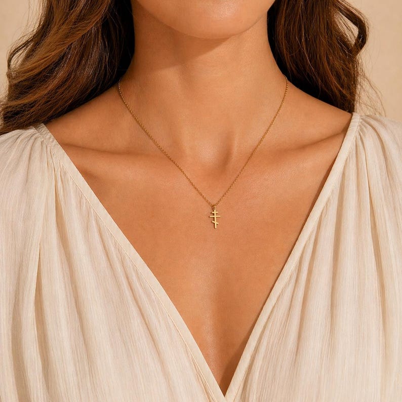 May include: A gold-colored necklace with a delicate pendant featuring a stacked cross design. The necklace is displayed on a person wearing a cream-colored blouse. The pendant is small and simple, adding a touch of elegance.