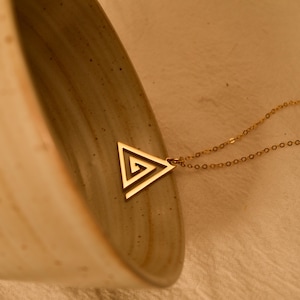 May include: A gold-toned necklace featuring a triangular pendant with an open-work spiral design. The pendant is attached to a delicate gold-toned chain with small circular links. The metal has a smooth, polished finish that reflects light.
