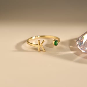 Op de afbeelding: Een gouden ring met een groene edelsteen en de letter "K" op de band.