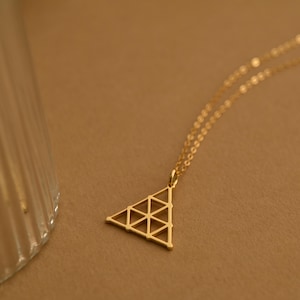 May include: A gold necklace featuring a geometric triangle pendant. The pendant has an open design with small triangles inside the larger shape. The chain is fine and the necklace is shown against a neutral backdrop.