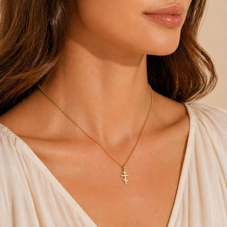 May include: A gold-colored necklace with a triple cross pendant. The delicate chain hangs on a person's neck, with the pendant resting near the chest. The person is wearing a white top.