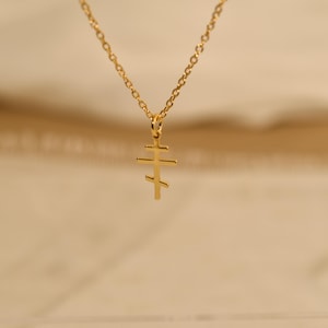 May include: A gold-colored necklace featuring a pendant in the shape of a Russian Orthodox cross. The cross has three horizontal bars, with the top bar being shorter than the middle and bottom bars. The chain is also gold and has a delicate link design.