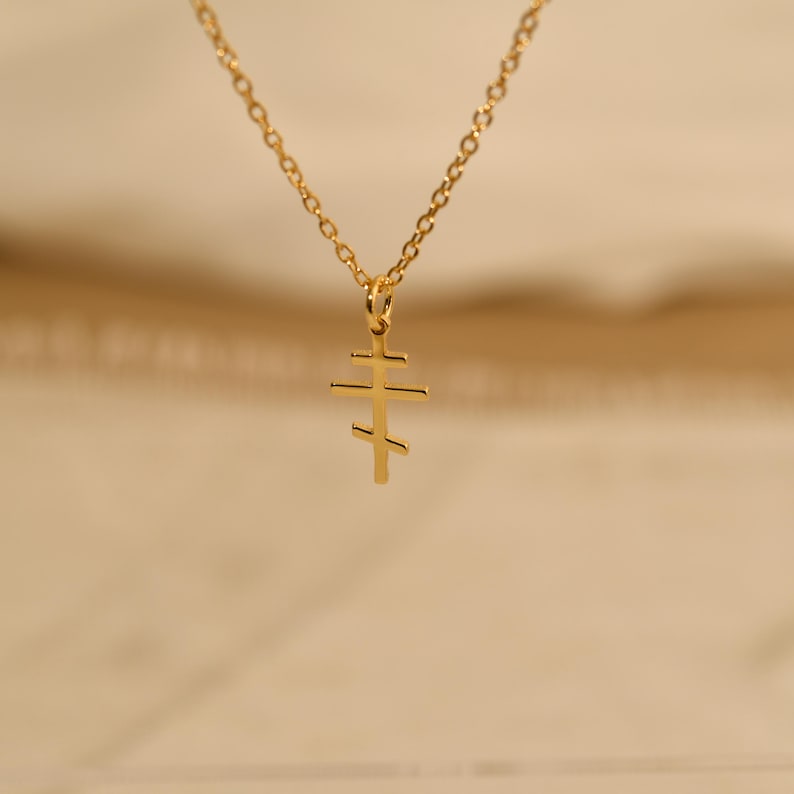 May include: A gold-colored necklace featuring a pendant in the shape of a Russian Orthodox cross. The cross has three horizontal bars, with the top bar being shorter than the middle and bottom bars. The chain is also gold and has a delicate link design.