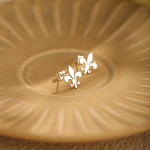 Op de afbeelding: Een paar zilverkleurige oorknopjes met een fleur-de-lis ontwerp. Elke oorbel heeft een gepolijste metalen afwerking en een standaard sluiting, waarbij het iconische symbool van de driebladige lelie in een strakke vorm wordt getoond.