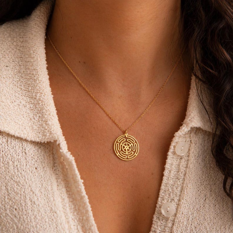 May include: A gold-colored pendant necklace featuring a circular design with a maze-like pattern. The pendant hangs from a delicate chain. The necklace is worn against a cream-colored shirt.