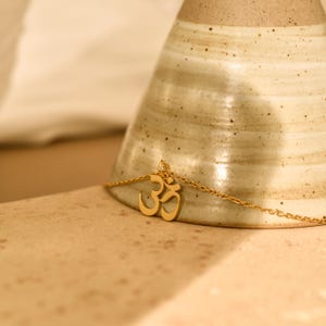 Puede incluir: Un collar dorado con un colgante con el símbolo Om. El colgante cuelga de una cadena fina. El collar se muestra sobre una superficie marrón claro, con un objeto de cerámica borroso en el fondo.