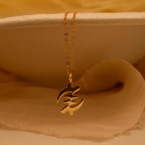 May include: A gold-toned necklace featuring a delicate chain and a pendant shaped like the Gye Nyame Adinkra symbol. The pendant has a polished, reflective surface and is suspended from a thin, matching gold-toned metal link chain.