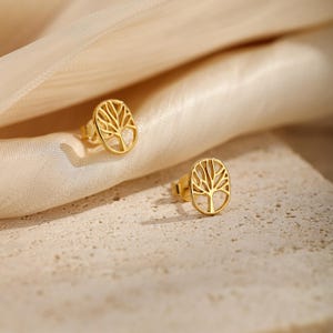 Puede incluir: Un par de pendientes de botón en tono dorado, cada uno con un marco circular que contiene un diseño recortado detallado de un árbol con ramas extendidas y un tronco. Los pendientes tienen un acabado metálico pulido y cierres de presión.