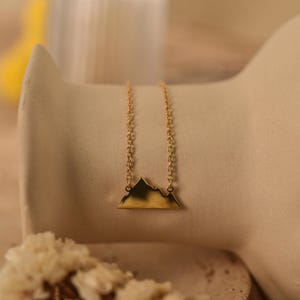 May include: A gold chain necklace with a mountain range pendant. The pendant is a simple silhouette of a mountain range.