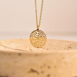 May include: A gold-colored pendant necklace featuring a circular labyrinth design. The pendant hangs from a delicate chain. The labyrinth design has a central star-like shape. The necklace is displayed on a textured, light-colored surface.
