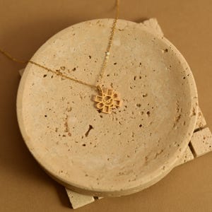 May include: A gold-colored necklace with a pendant in the shape of a stylized flower or sun. The necklace is displayed on a round, textured, beige stone dish. The chain is delicate, and the pendant has an open, geometric design.