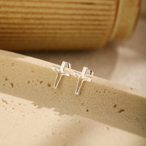 Könnte beinhalten: Ein Paar silberfarbene Ohrstecker in Form von minimalistischen Kreuzen. Jeder Ohrstecker besteht aus einem schlanken, länglichen vertikalen Balken, der von einem kürzeren horizontalen Balken gekreuzt wird, was eine klare, polierte Metall-Silhouette ergibt.