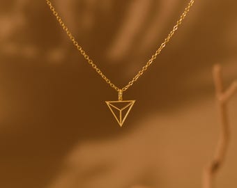 May include: A gold-colored necklace with a geometric triangle pendant. The pendant is a three-dimensional open triangle design. The chain is delicate and the necklace is displayed against a warm, neutral background.