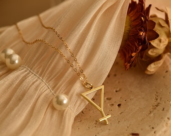 May include: A gold-colored necklace with a pendant shaped like a stylized female symbol. The necklace is on a delicate gold chain, and the pendant has a triangular top and a cross at the bottom. The image also shows several white pearls and a dried flower.