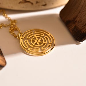 May include: A gold-colored pendant necklace featuring a circular design with a maze-like pattern and a star-shaped center. The necklace is on a delicate chain, and the pendant is the focal point of the image. The background is a soft, neutral color.
