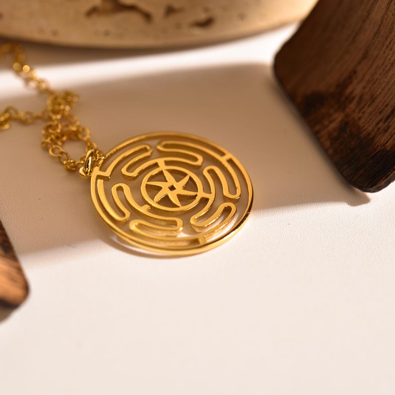 May include: A gold-colored pendant necklace featuring a circular design with a maze-like pattern and a star-shaped center. The necklace is on a delicate chain, and the pendant is the focal point of the image. The background is a soft, neutral color.
