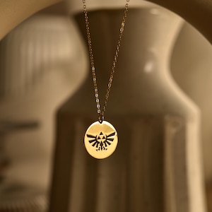 Pode incluir: Um colar com pingente dourado com um berloque circular com um emblema preto. O colar tem uma corrente delicada e é suspenso contra um fundo desfocado. O design do pingente é um emblema estilizado.