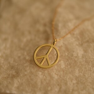 May include: A gold-colored necklace featuring a peace symbol pendant. The pendant is a circular outline with a vertical line and two diagonal lines inside. The chain is delicate and gold-toned. The necklace is displayed on a textured, light-colored surface.