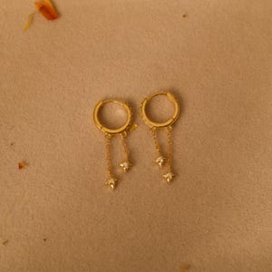 May include: Gold-coloured hoop earrings with star charms. The hoops are decorated with small, clear stones. Each earring has a chain with a star-shaped charm, creating a celestial design. The earrings are displayed on a neutral background.