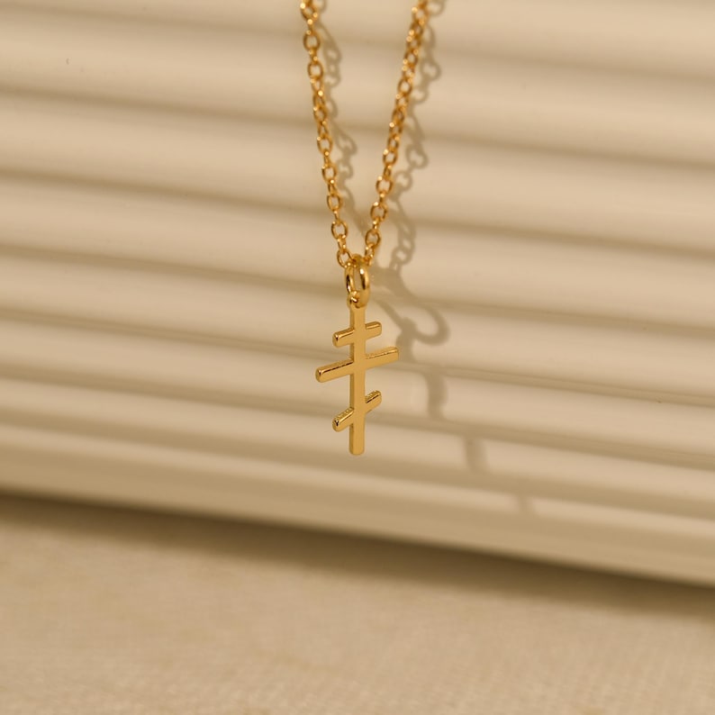 May include: A gold-colored necklace with a pendant in the shape of a cross. The cross has three horizontal bars. The chain is delicate and the pendant is small. The necklace is displayed against a neutral background.