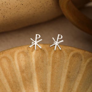 May include: A pair of silver-colored stud earrings featuring the Chi Rho symbol. Each earring consists of a vertical line crossed by a diagonal line, with a loop at the top of the vertical stem, forming the Greek letters Chi and Rho in a metallic finish.