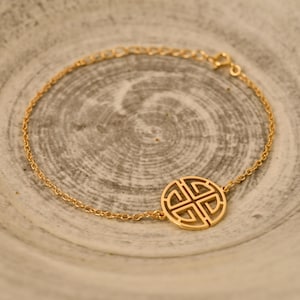 May include: A gold-colored bracelet with a delicate chain and a circular pendant. The pendant features an intricate, openwork design. The bracelet is displayed on a neutral, textured surface.