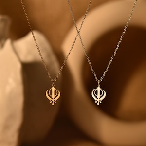 May include: Two gold necklaces with a Khanda pendant. The necklaces are on a light brown background.