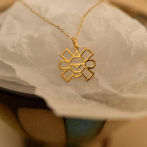 May include: A gold-colored pendant necklace featuring a geometric design. The pendant has a sun-like shape with a central eye-like detail. The necklace is displayed on a white, textured surface.