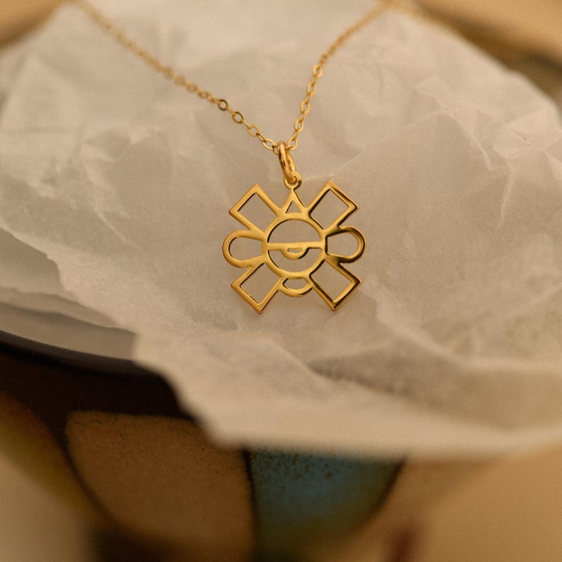 May include: A gold-colored pendant necklace featuring a geometric design. The pendant has a sun-like shape with a central eye-like detail. The necklace is displayed on a white, textured surface.