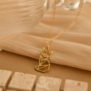 Puede incluir: Un collar dorado con un colgante geométrico de zorro. El colgante está hecho de líneas interconectadas que forman la forma de un zorro. La fina cadena cuelga sobre un fondo de tela beige. La joya se exhibe en un estudio.