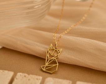 Puede incluir: Un collar dorado con un colgante geométrico de zorro. El colgante está hecho de líneas interconectadas que forman la forma de un zorro. La fina cadena cuelga sobre un fondo de tela beige. La joya se exhibe en un estudio.