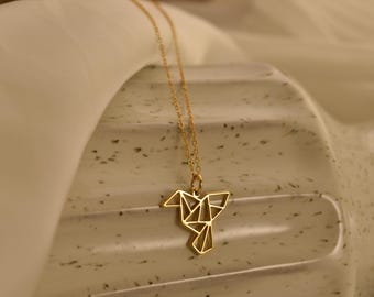 May include: A gold-colored necklace with a geometric bird pendant. The pendant is a stylized origami design, suspended from a delicate chain. The necklace is displayed on a textured, light-colored surface.